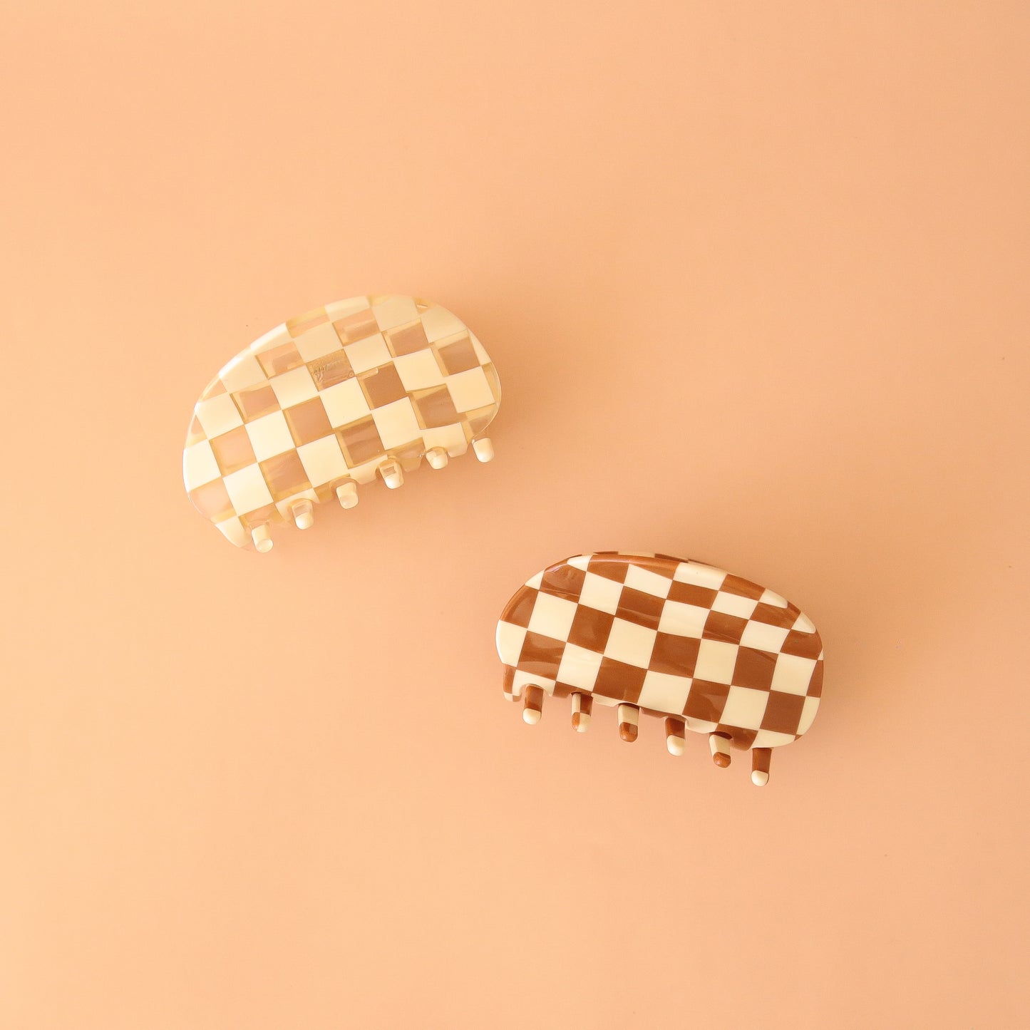 On a peachy background is two claw clips with a rounded edge design. One of the claw clips has an ivory and clear checker pattern while the other clip has a brown and ivory checker pattern.