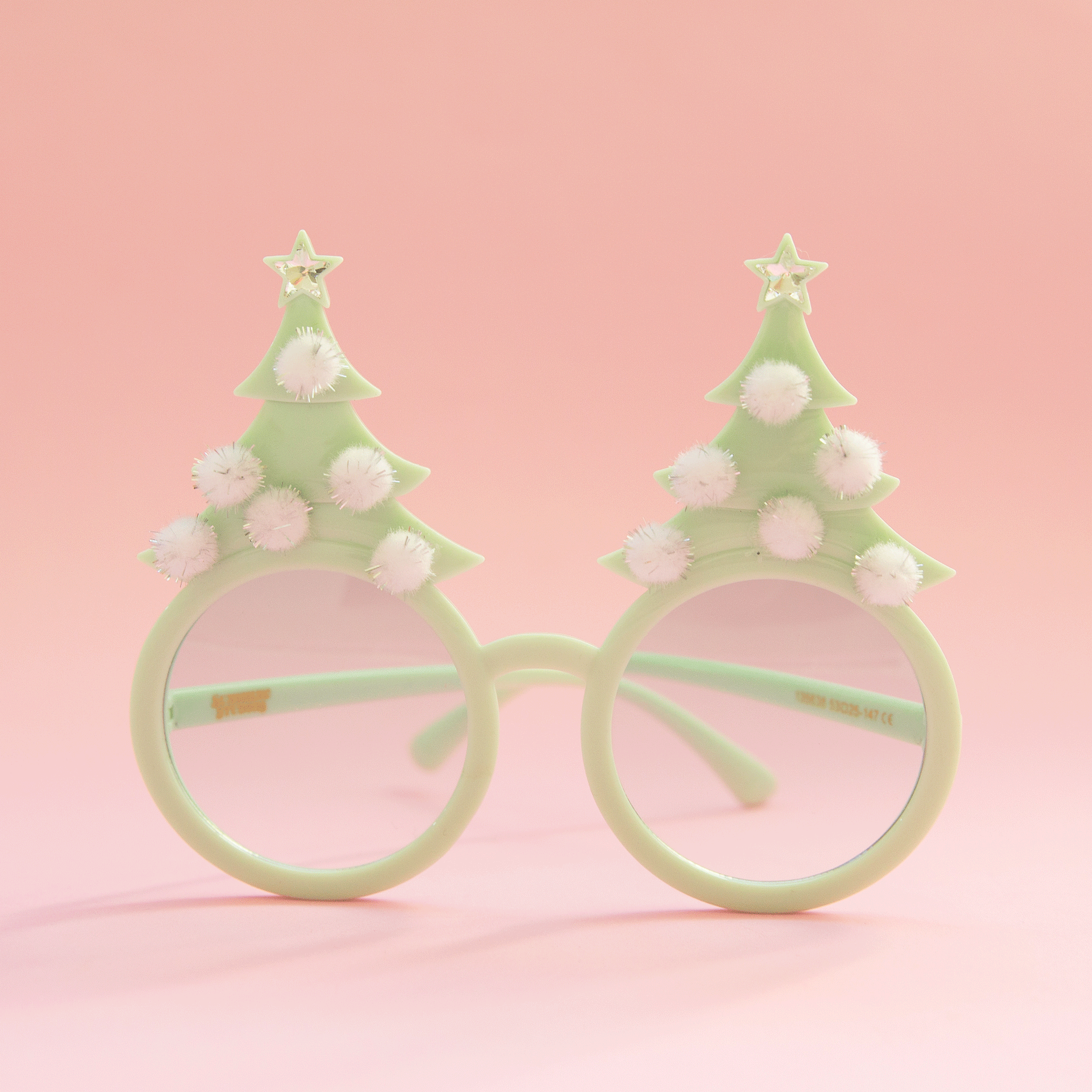 On a pink background is a pair of mint holiday glasses with tree shapes and six white pom poms and a star on top.