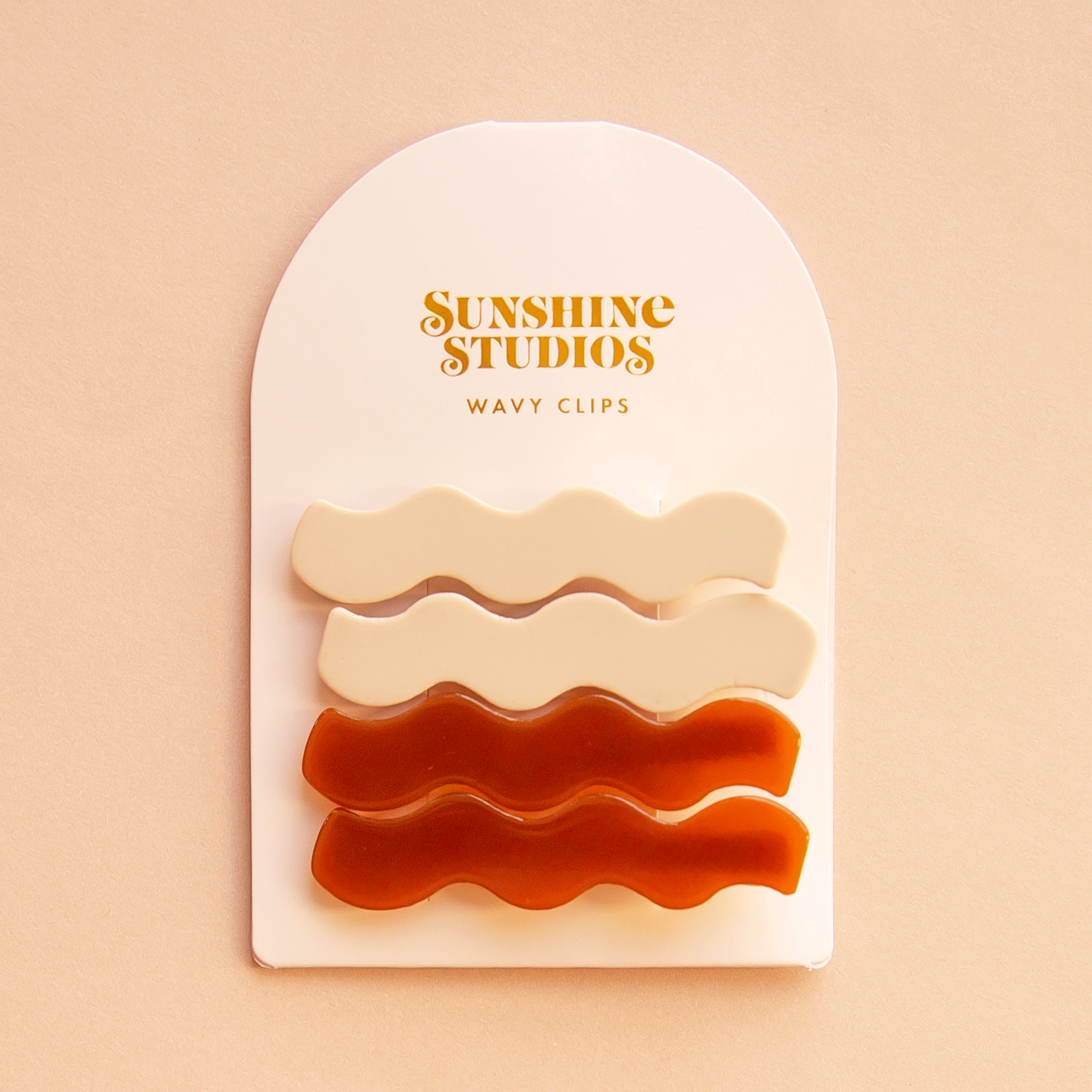 On a peach background is two pairs of wavy hair clips on a white arched cardboard backing with text at the top that reads, "Sunshine Studios Wavy Clips".