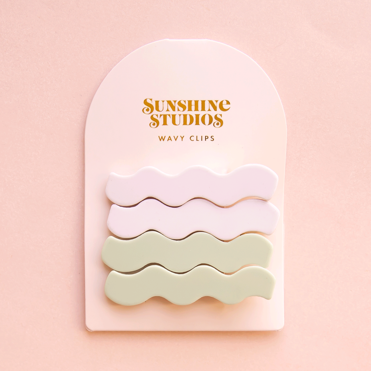 On a pink background is two sets of wavy shaped hair clips in a light green/blue shade and shade of white on an arched packaging with text along the top that reads, "Sunshine Studios".