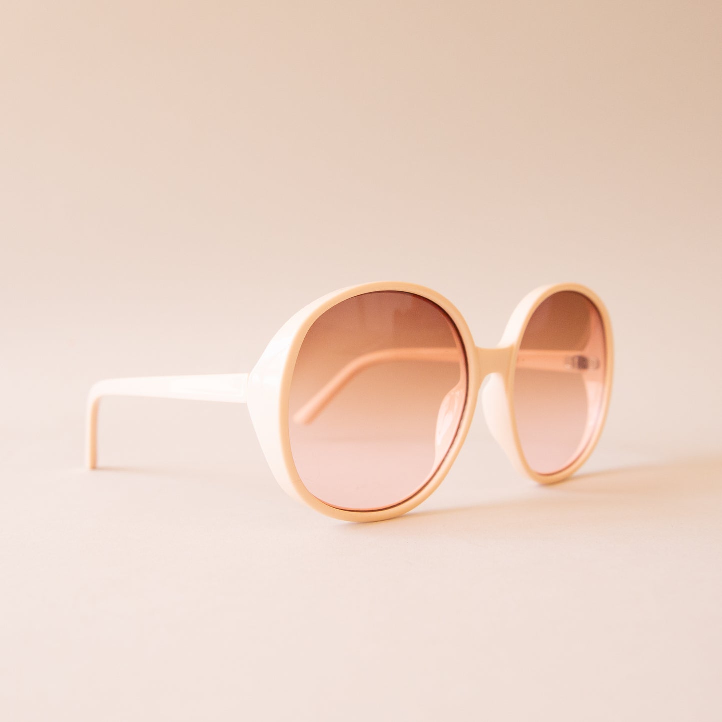 A creamy white pair of round oversized sunglasses with a light brown / pink lens.