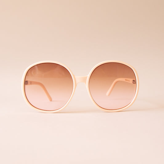 A creamy white pair of round oversized sunglasses with a light brown / pink lens. 