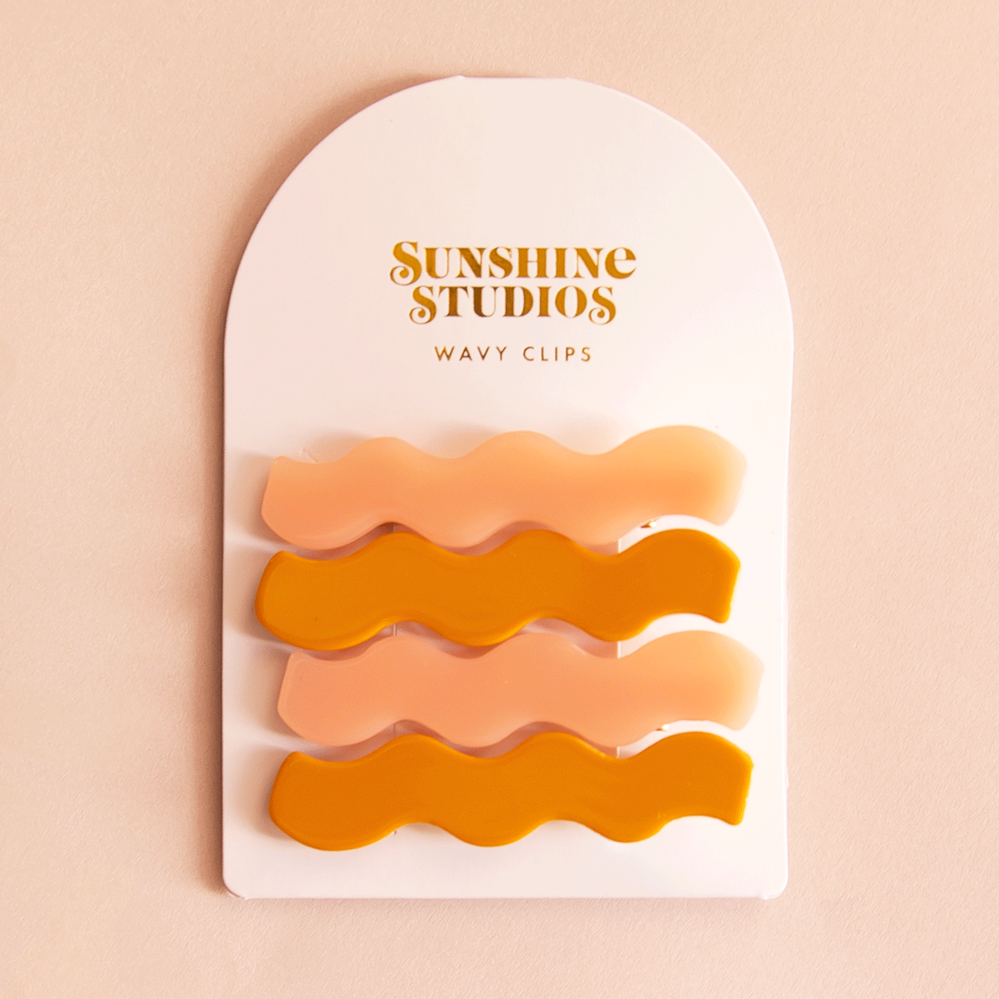 On a pink background two sets of wavy hair clips in a mango orange shade and a light pink shade clipped on a white arched piece of cardboard packaging with orange text at the top that reads, "Sunshine Studios".