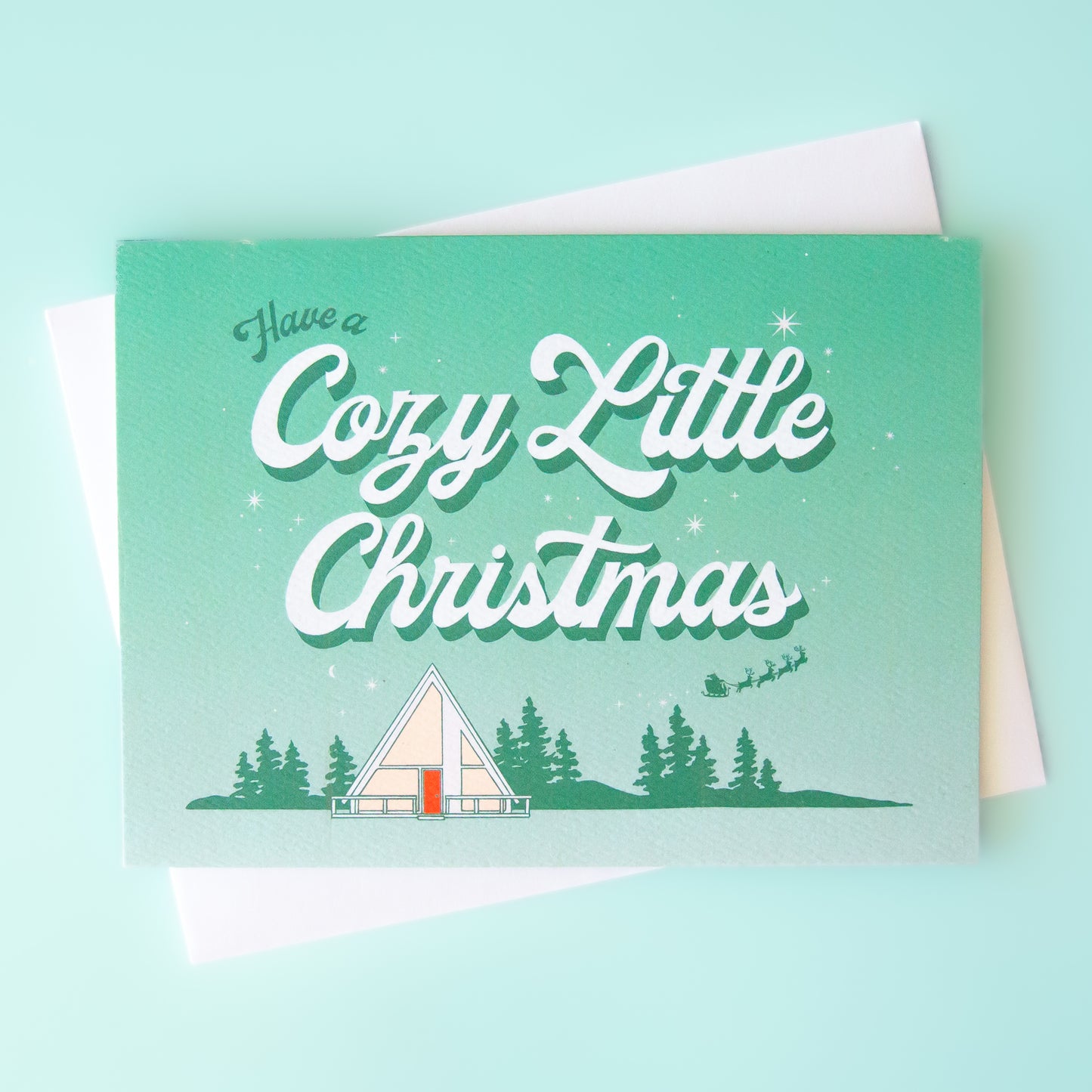 Aqua and white ombre greeting card that reads 'have a cozy little Christmas'. Below the text is a small white tent in a forest scene. A small Santa sleigh and reindeer fly across the sky.  The card is accompanied by a white envelope. 