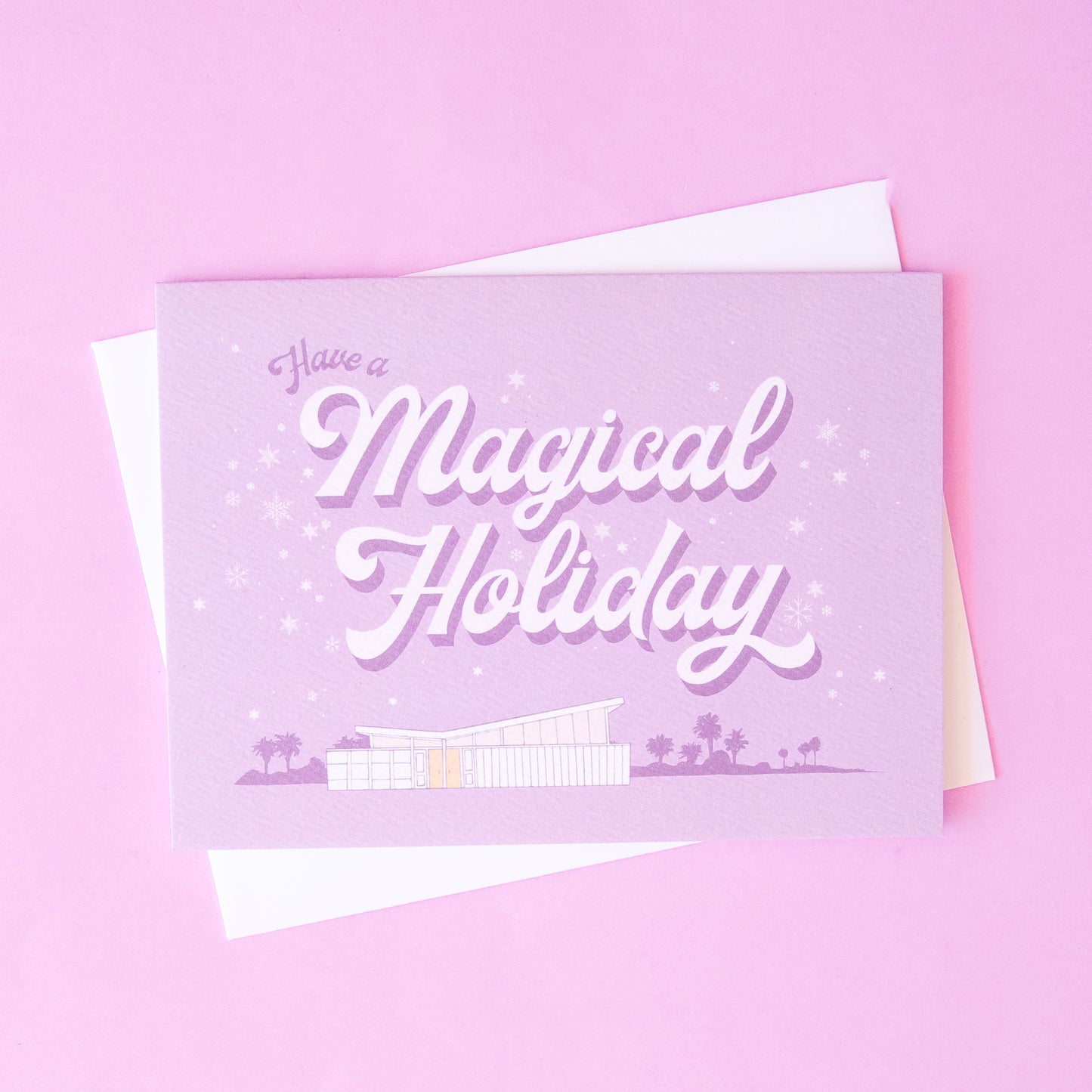 Lilac holiday card that reads 'have a a magical holiday' in cursive lettering. Around the text is delicate white snowflakes. Below is a coastal scene of a beach house and palm trees.