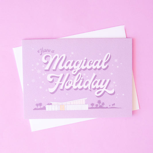 Lilac holiday card that reads 'have a a magical holiday' in cursive lettering. Around the text is delicate white snowflakes. Below is a coastal scene of a beach house and palm trees.