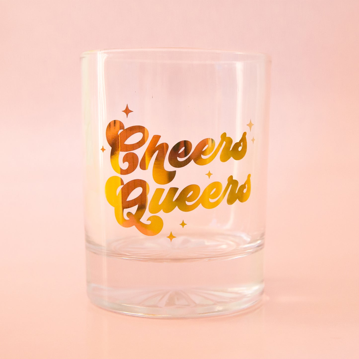 Glass tumbler with gold foil writing, spelling"cheers queers" is shown against a light pink background