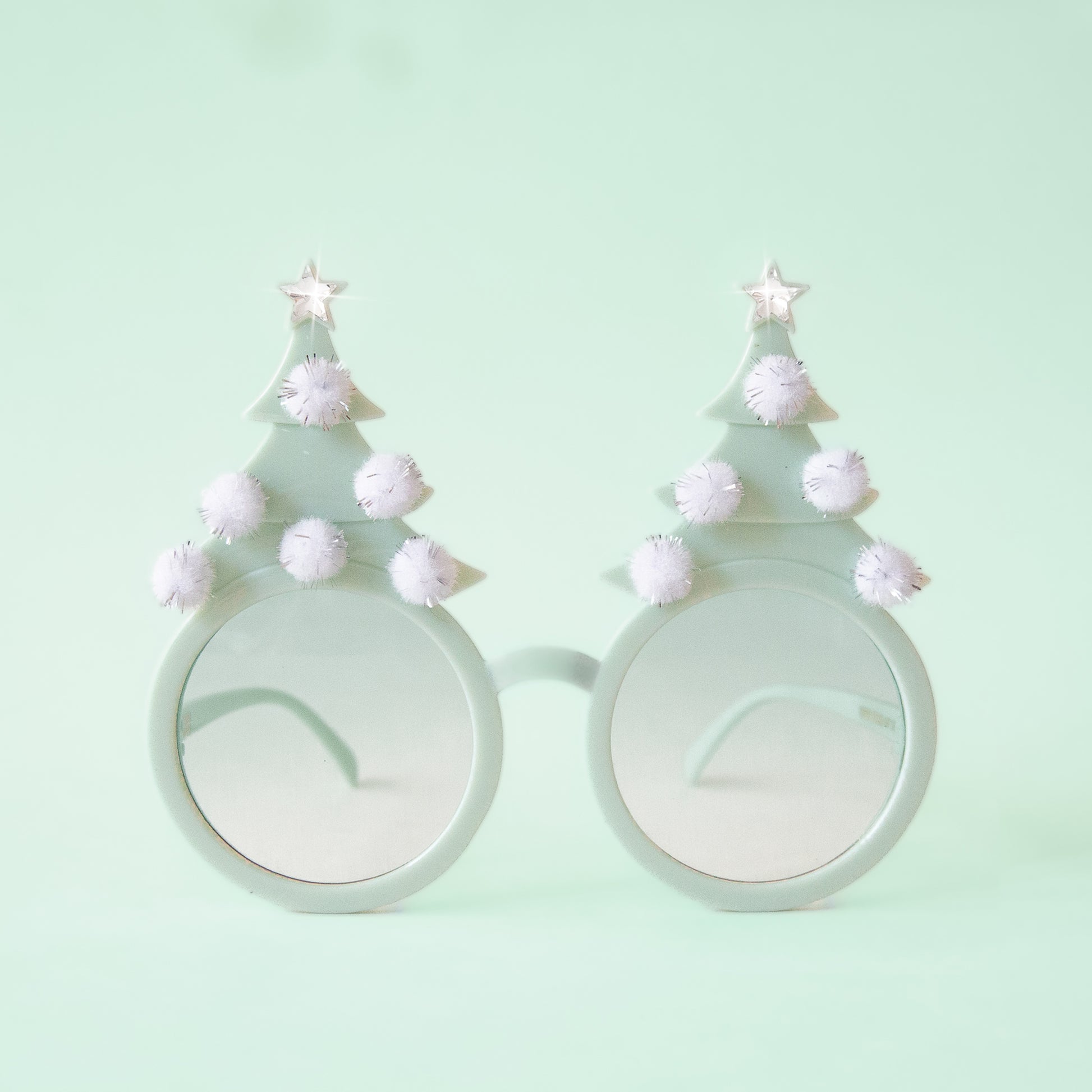 On a mint background is a pair of mint holiday glasses with tree shapes and six white pom poms and a star on top.