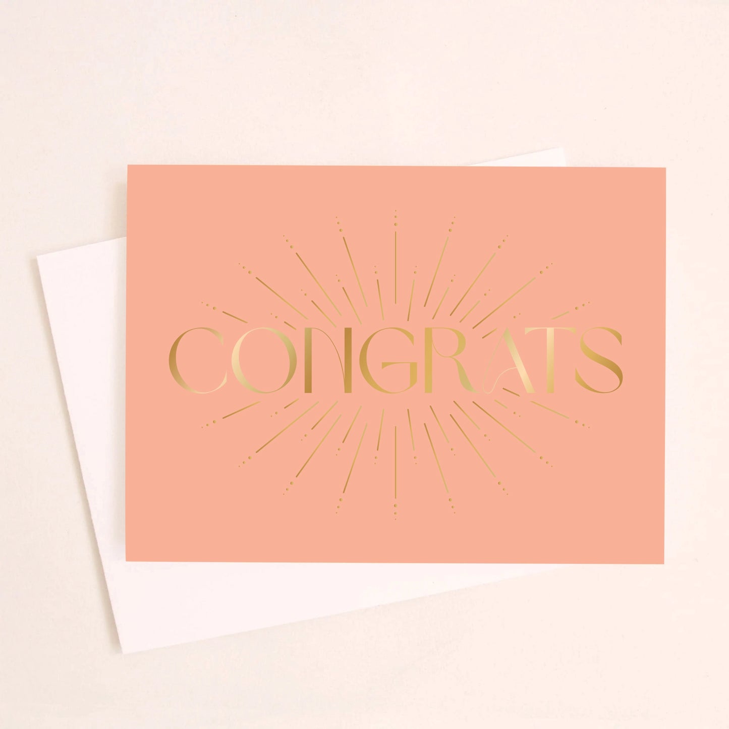 On an ivory background is a salmon colored greeting card with gold foil text that reads, "Congrats" along with a white envelope.