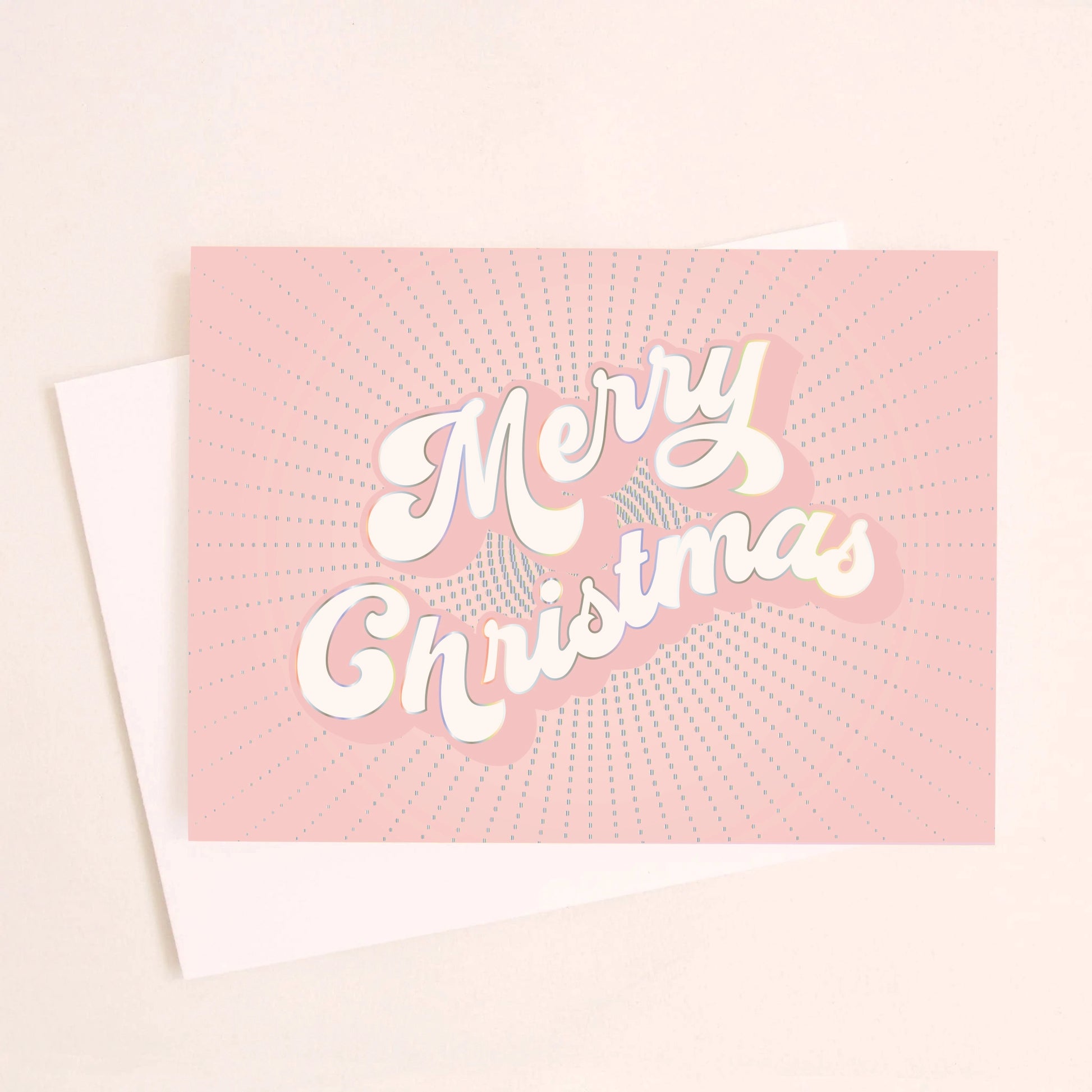 On a light pink background is a light pink greeting card with white text in the center that reads, "Merry Christmas" along with a white envelope. 