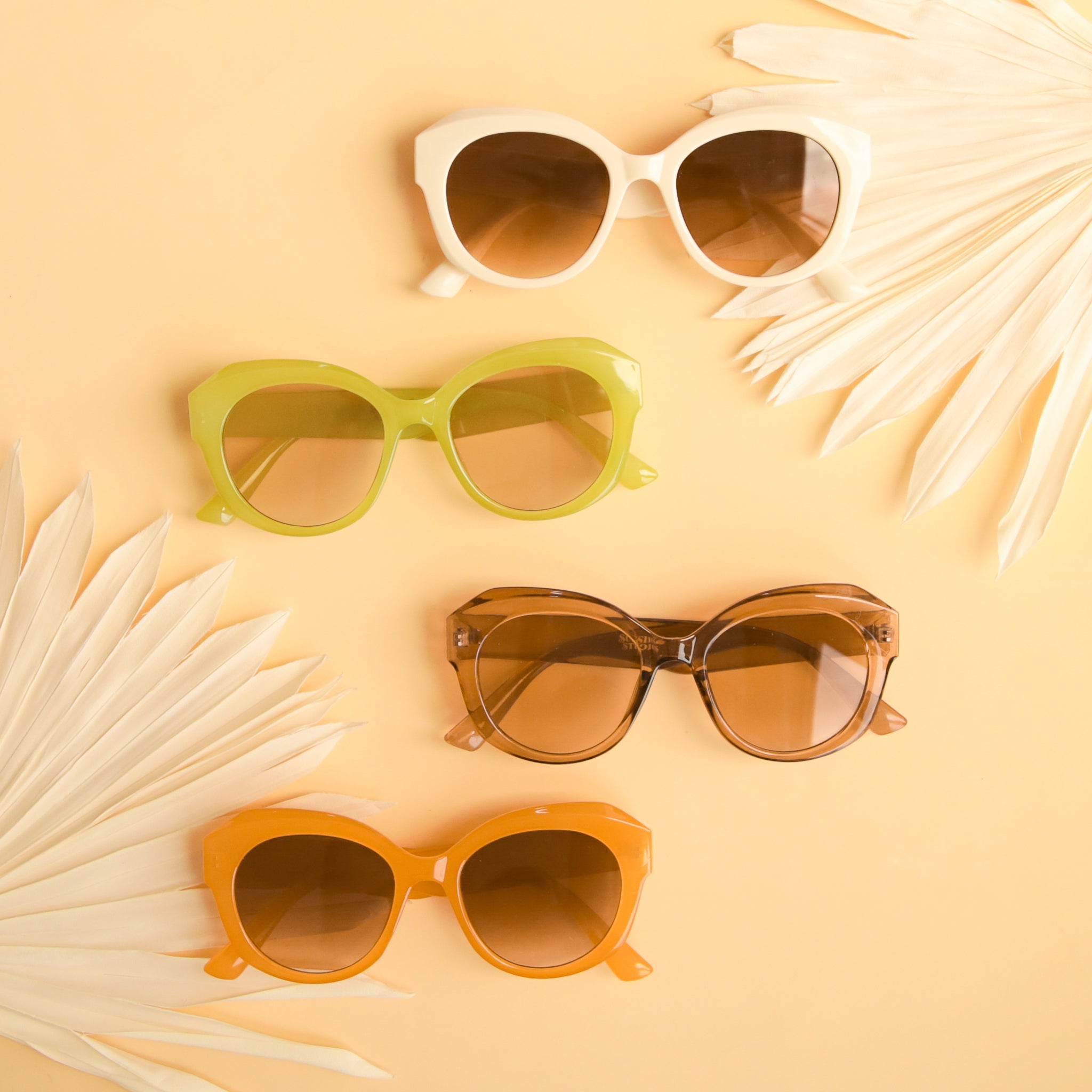 four pairs of retro styled sunglasses in multiple colors lies on a soft yellow ground with white dried palms