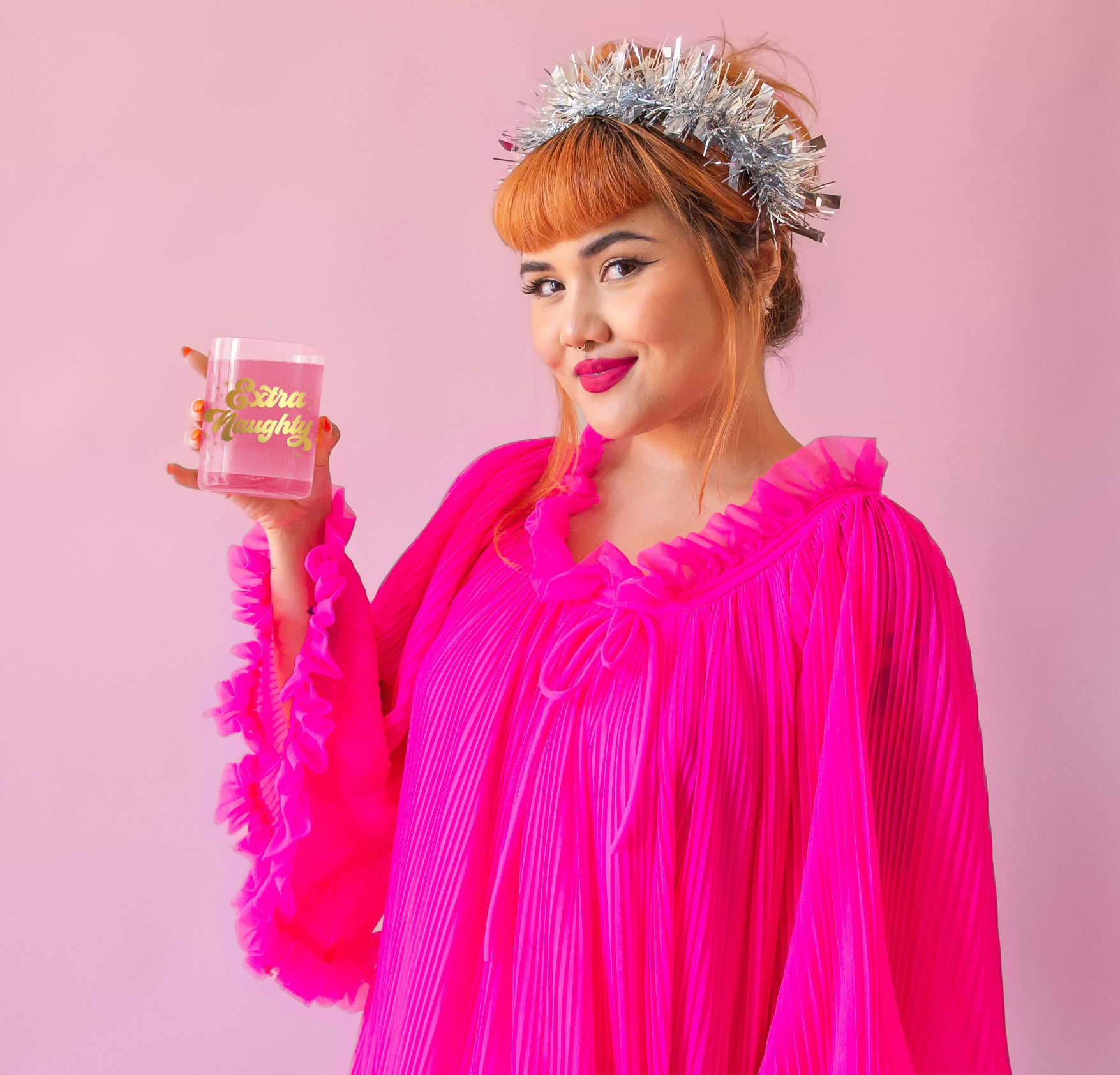 Model is holiday a clear glass with gold writing, wearing a bright pink dress and a festive silver headband 