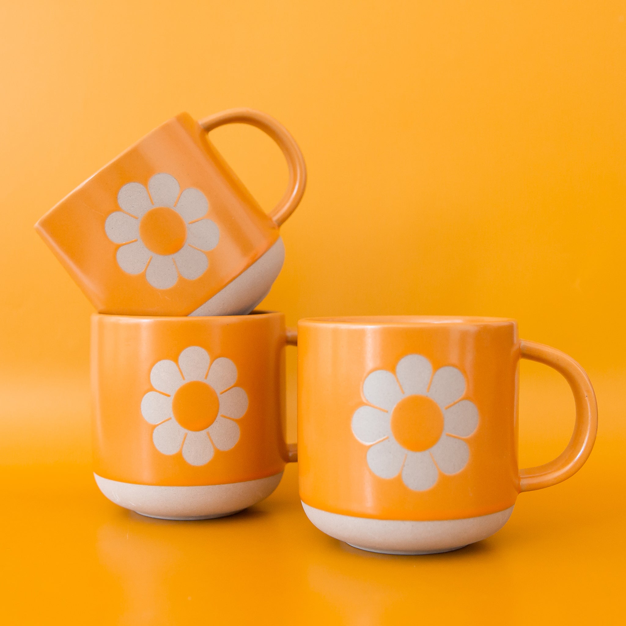 three mustard colored mugs with a daisy on the front sit stacked on a yellow-orange ground