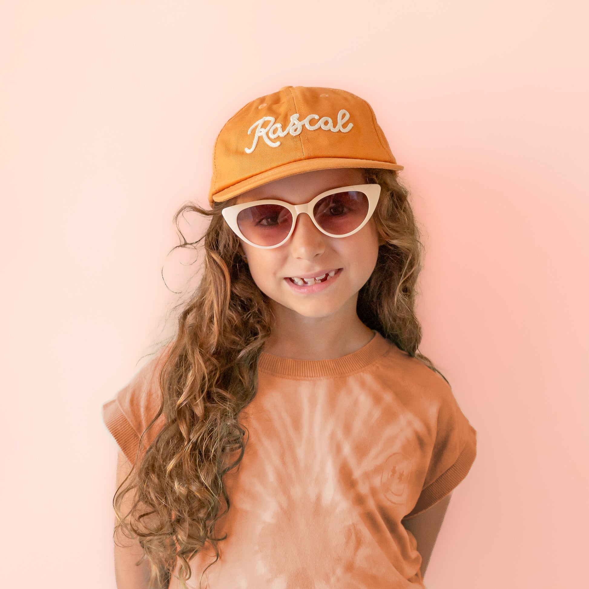 A burnt orange flat brimmed hat with white embroidered text that reads, "Rascal".