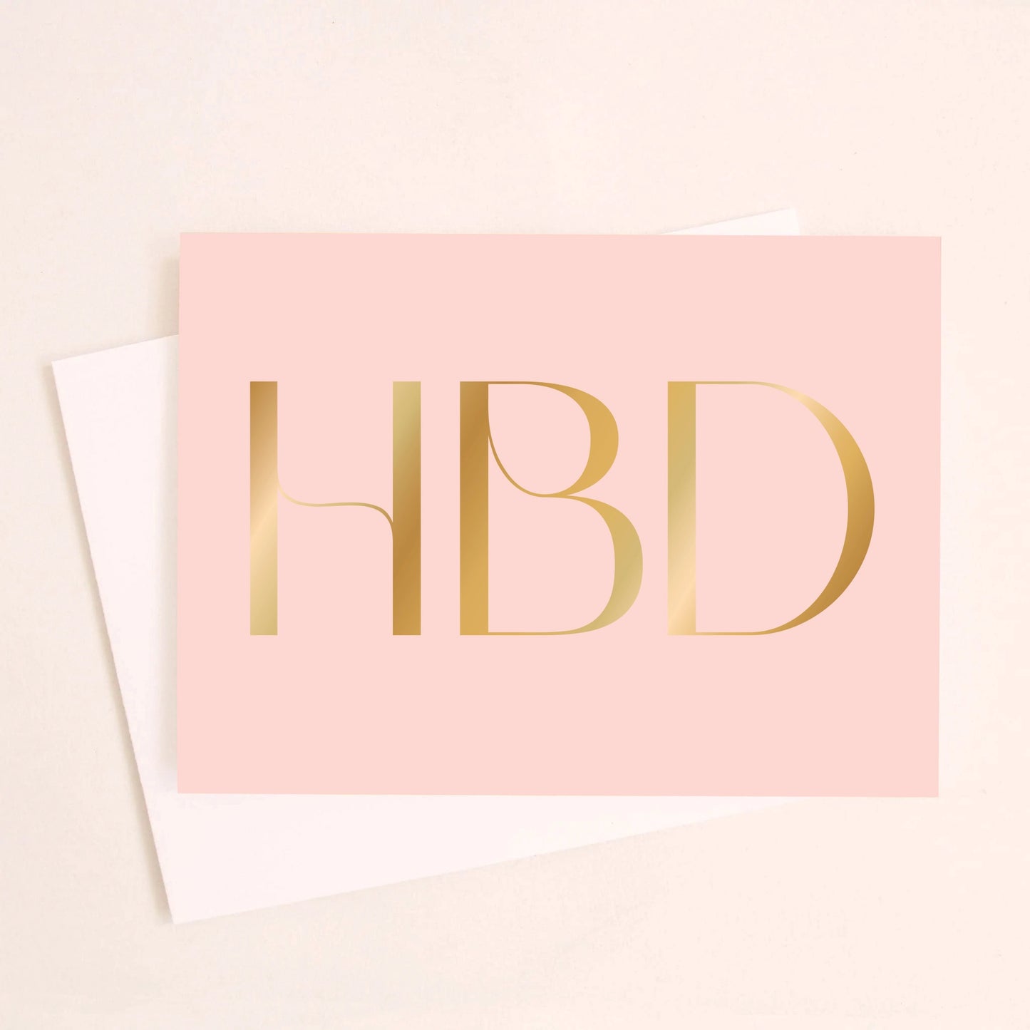 On an ivory background is a light pink greeting card with large gold foil text in the center that reads, "HBD" in all caps. Also included is a white envelope. 