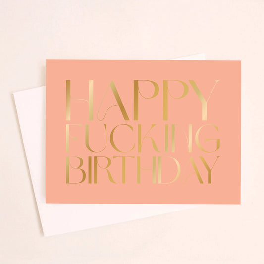 On an ivory background is a salmon colored greeting card with gold foil text in the center that reads, "Happy Fucking Birthday". 