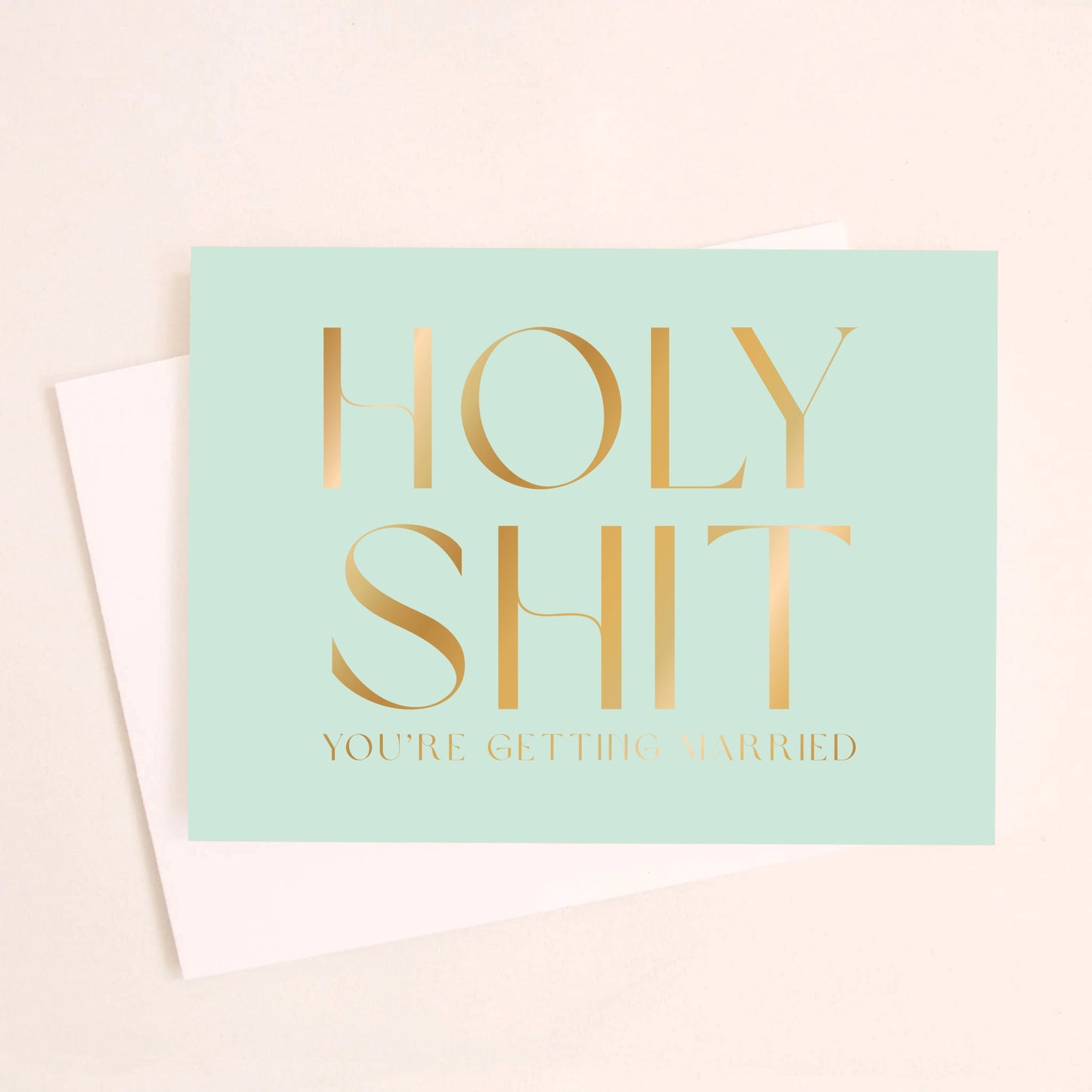 On an ivory background is a teal greeting card that reads, "Holy Shit You're Getting Married" in gold foil letters and a matching envelope beside it. 