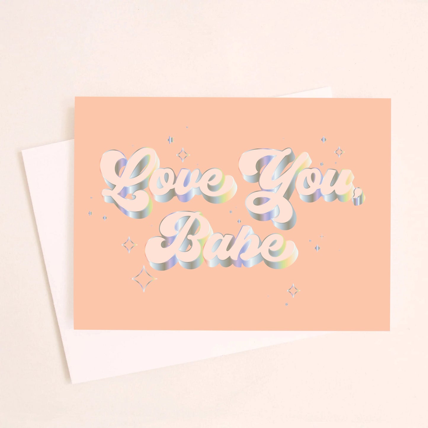 On a light peach background is a peachy greeting card with "Love You Babe" text in the center that is outlined with holographic foil detailing. Also included is a white envelope. 