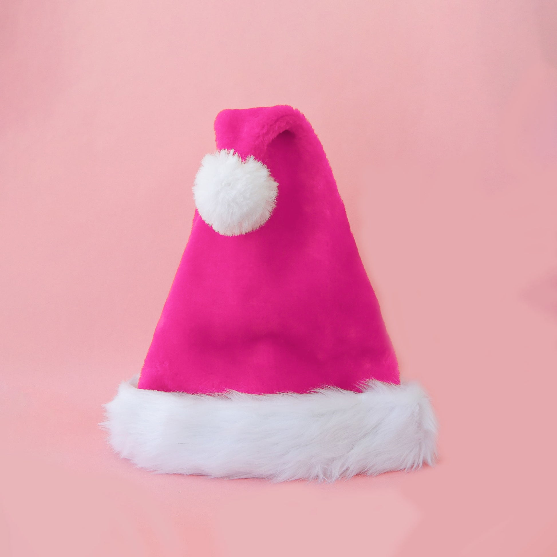 A hot pink faux fur Santa hat with white faux fur trim and pompom is shown against a light pink background.