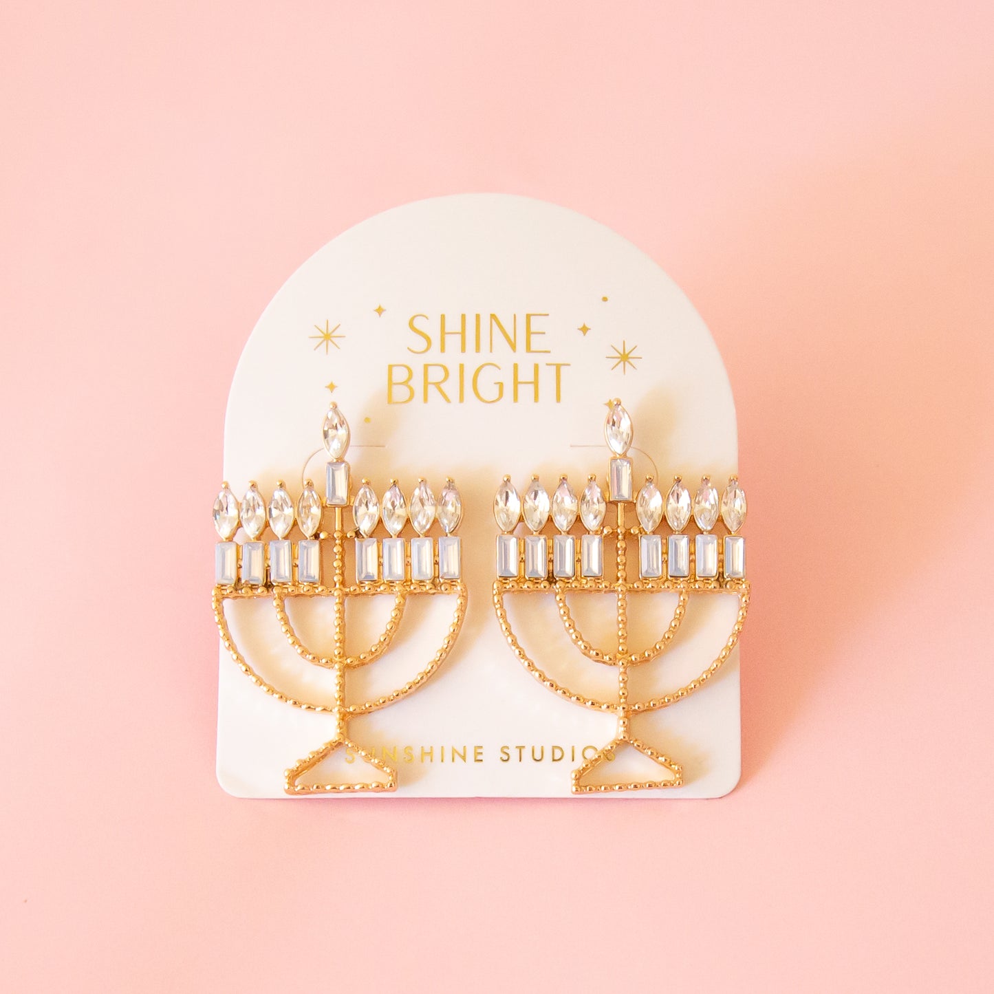 Menorah shaped gold earrings are attached to a cream colored card. the Card has gold foil stamp which reads Shine Bright and Sunshine Studios with glittering stars surrounding the text. The background of the photo is a soft warm pink color. 