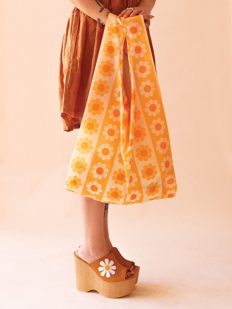 a woman in a brown dress holds an orange and yellow daisy reusable bag at her side