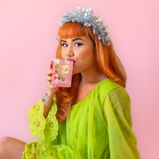 Model is sipping out of a glass cup with gold writing, wearing a festive silver headband and a bright green dress. 