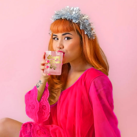 Model is sipping out of a glass cup with gold writing, wearing a festive silver headband and a bright pink dress. 