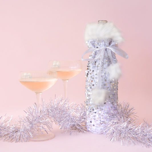 On a pink background is a silver sequin wine gift bag with white faux fur around the opening and a white pom pom bow around the top.