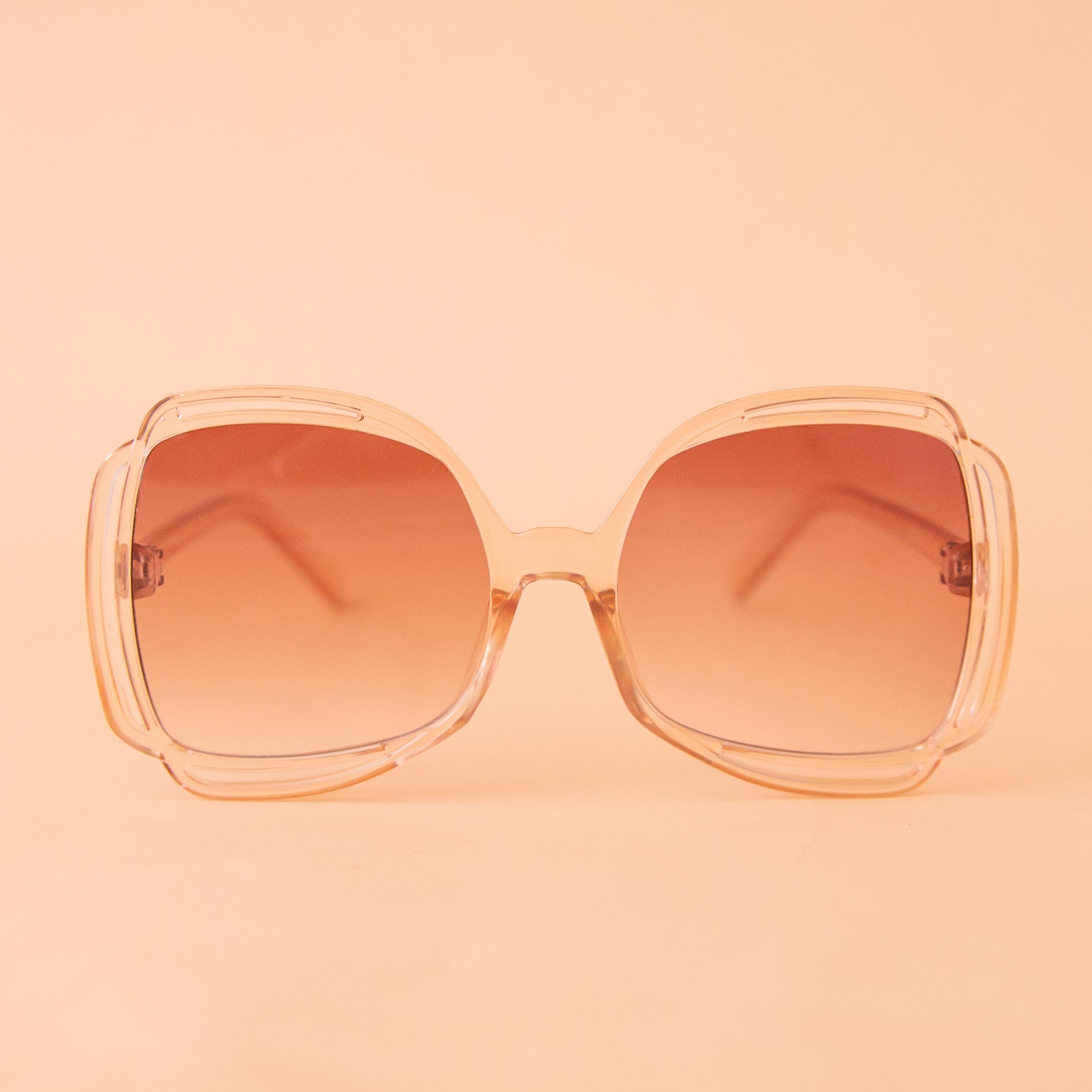 A pair of sunglasses with a clear peachy frame and pinkish gradient lenses. 