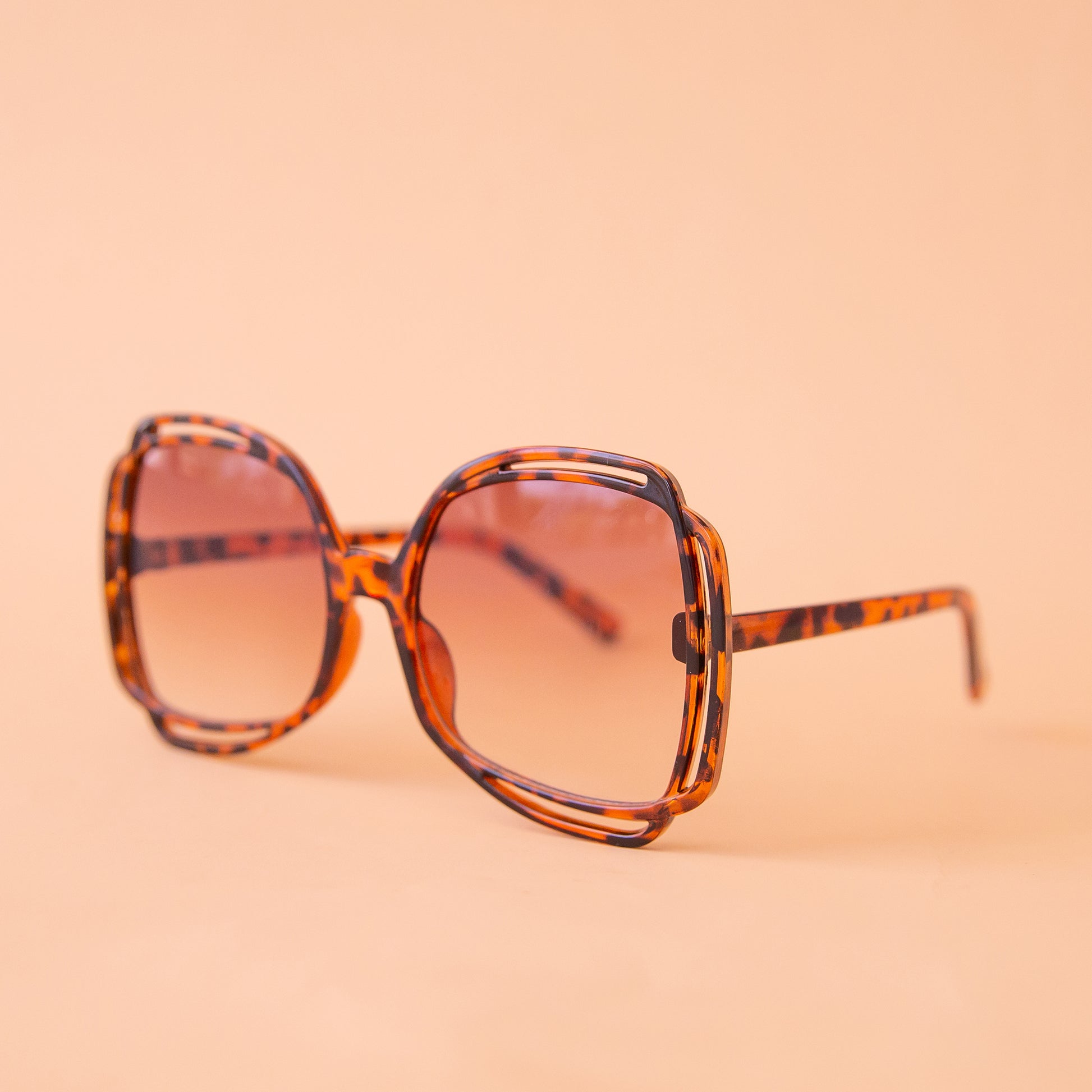 On a peachy background is a pair of tortoise sunglasses with a round frame and a brownish pink lens.