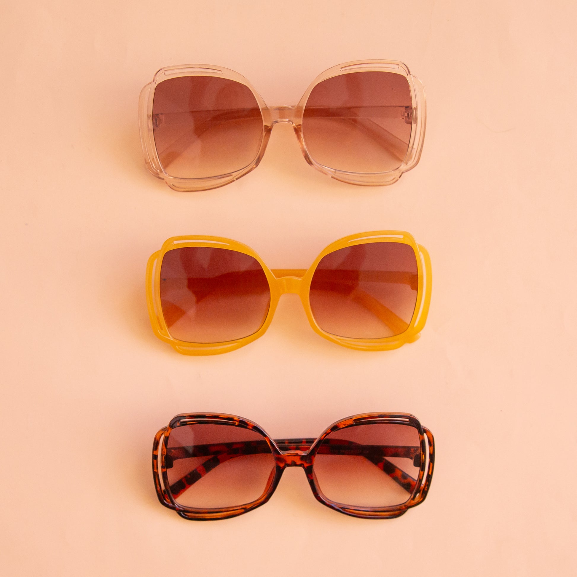 A pair of sunglasses with a clear peachy frame and pinkish gradient lenses sitting next to the other two available color options.