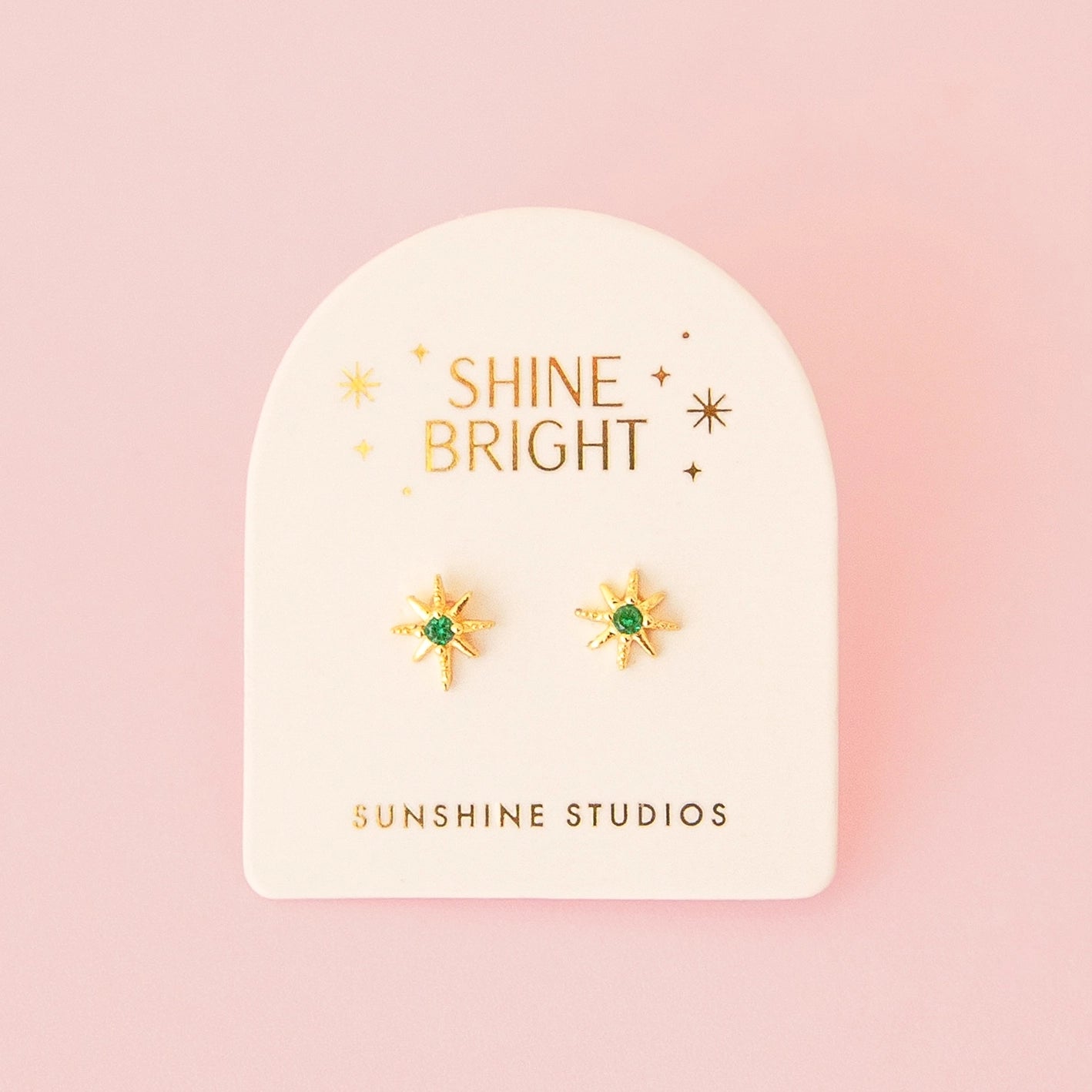 Gold star earring studs with a green gem in the center of the earrings. The earrings are on a paper backer. 