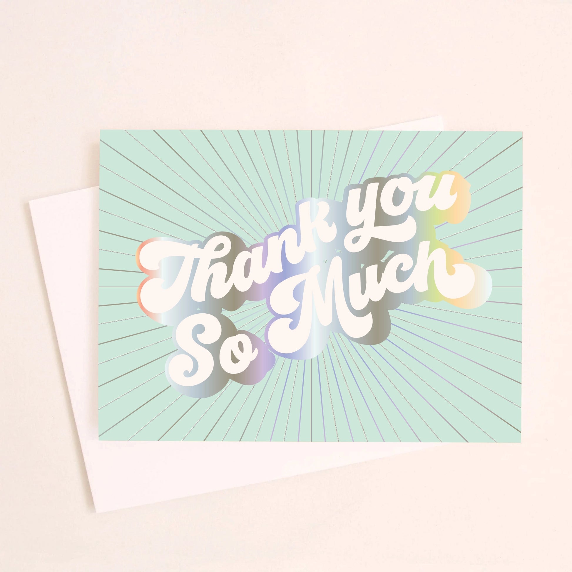 On an ivory background is a light teal greeting card with white, holographic outlined text that reads, "Thank You So Much". 