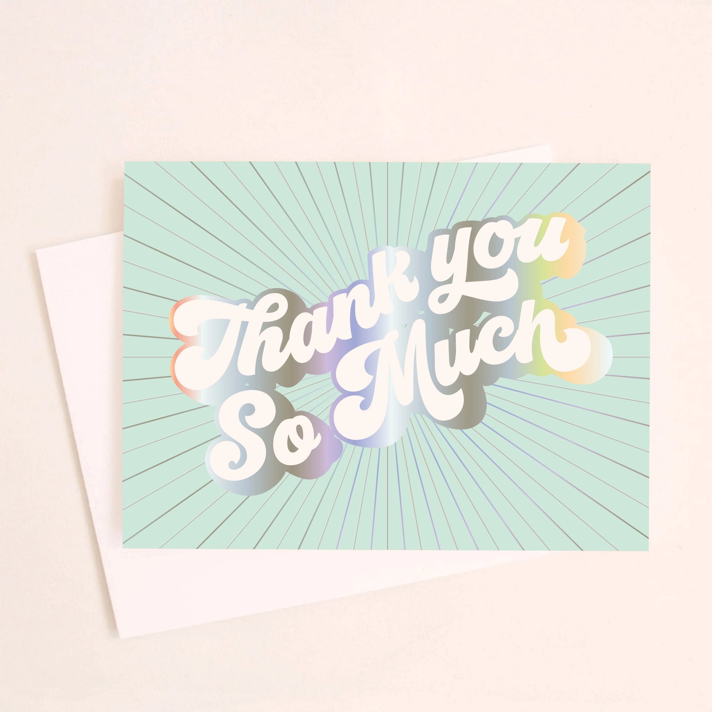 On an ivory background is a light teal greeting card with white, holographic outlined text that reads, "Thank You So Much". 