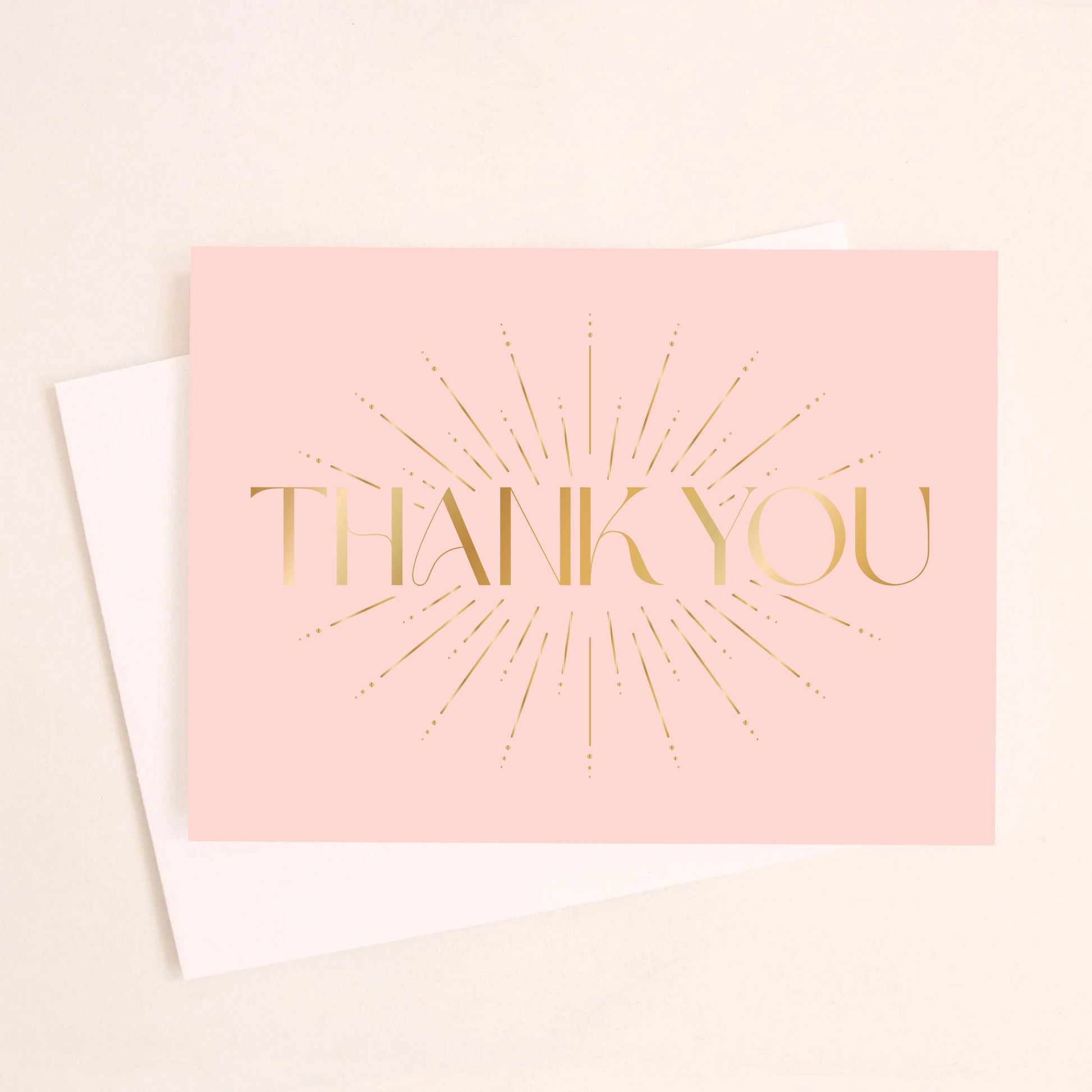 On an ivory background is a light pink card with a gold foil starburst design and text in the center that reads, "Thank You".
