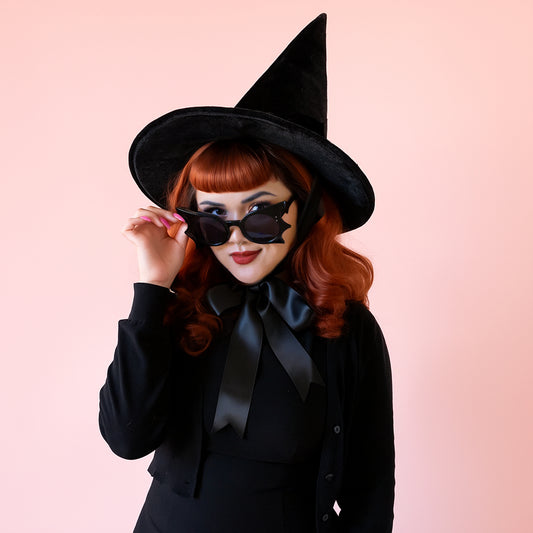 A red headed woman wears black bat shaped sunglasses and a tall black witch hat with a bow at the neck.  She is wearing an all black outfit.