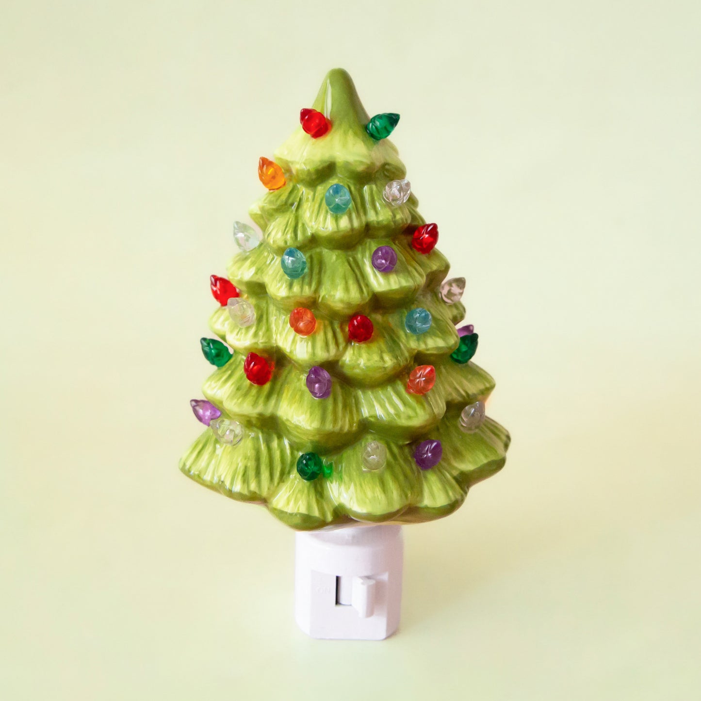 A ceramic Christmas Tree shaped night light sits in middle of the page with bright colorful lights.  It has a white plug at the bottom.  The tree is a lime green color.