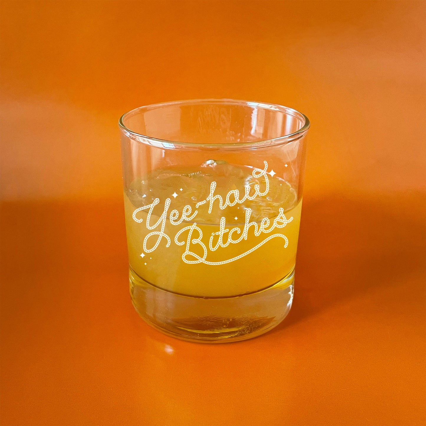 On an orange background is a short glass tumbler with white screenprinted text that reads, "Yee-haw Bitches". 