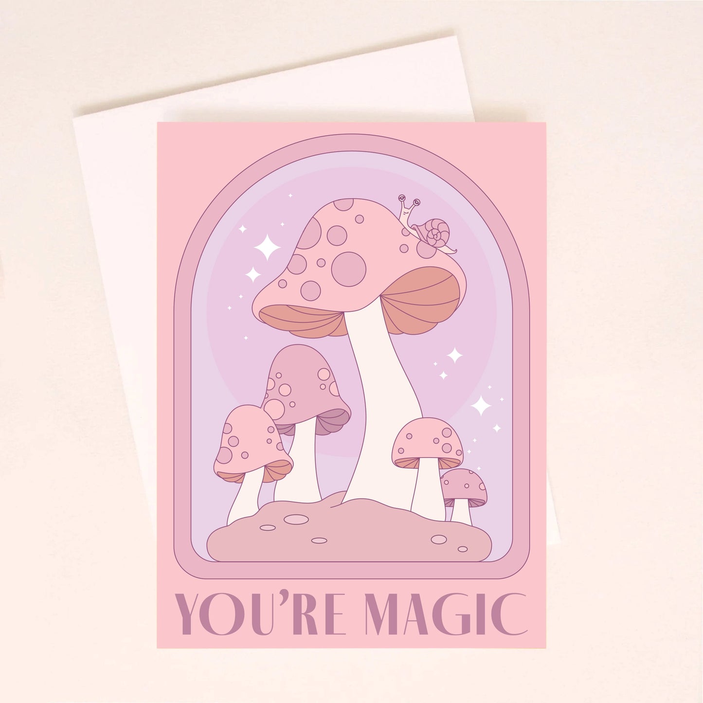 On a light pink background is a pink and purple card with an arch and mushroom design as well as text along the bottom that reads, "You're Magic". A white envelope is included with purchase, also shown here. 