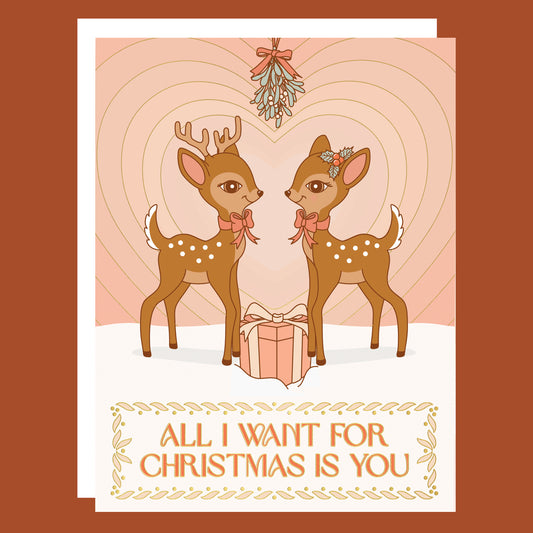 On a neutral background is a light pink card with two brown retro deer looking towards one another underneath mistletoe with a gradient heart in the background and text below the image that reads, "All I Want For Christmas Is You"
