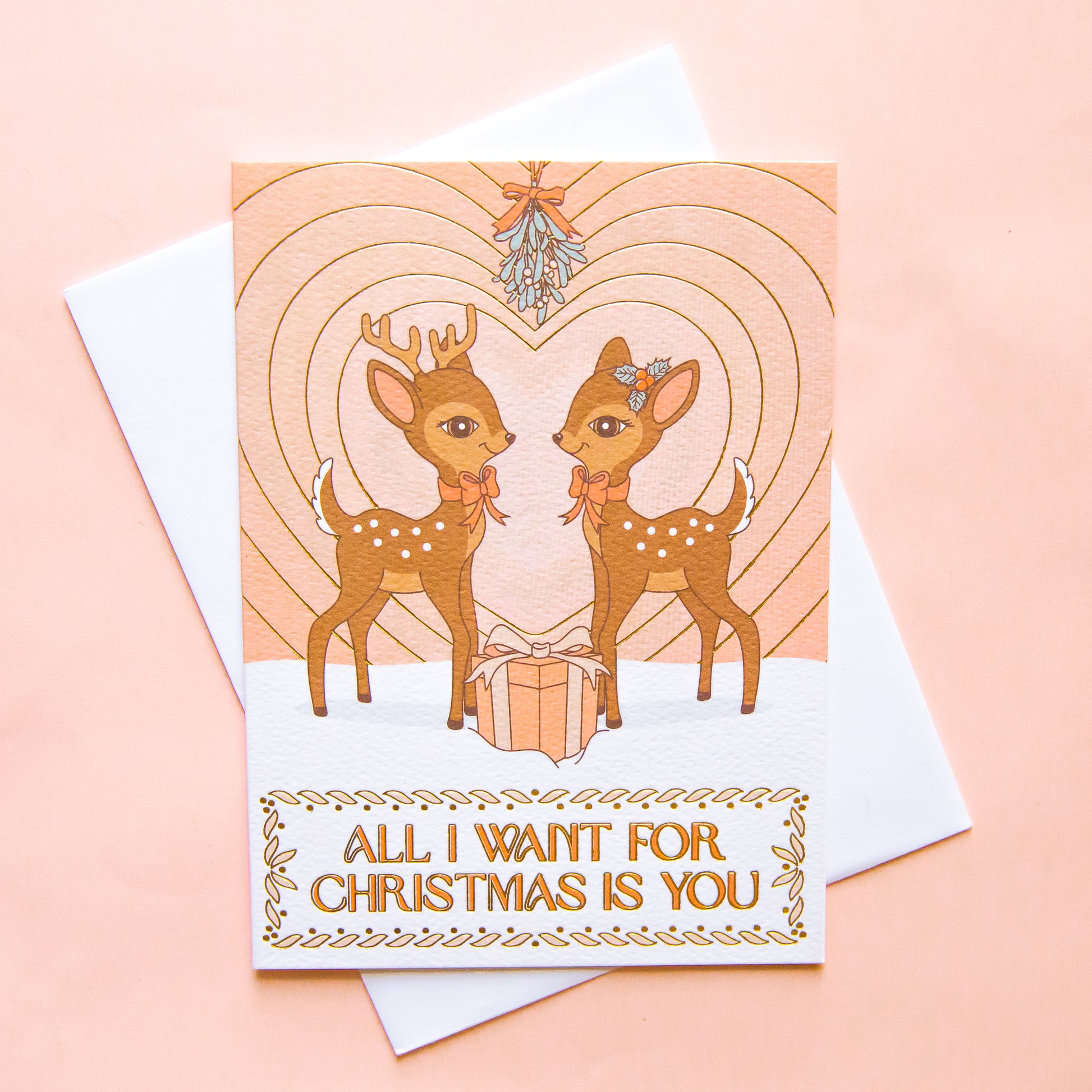 On a neutral background is a light pink card with two brown retro deer looking towards one another underneath mistletoe with a gradient heart in the background and text below the image that reads, "All I Want For Christmas Is You".