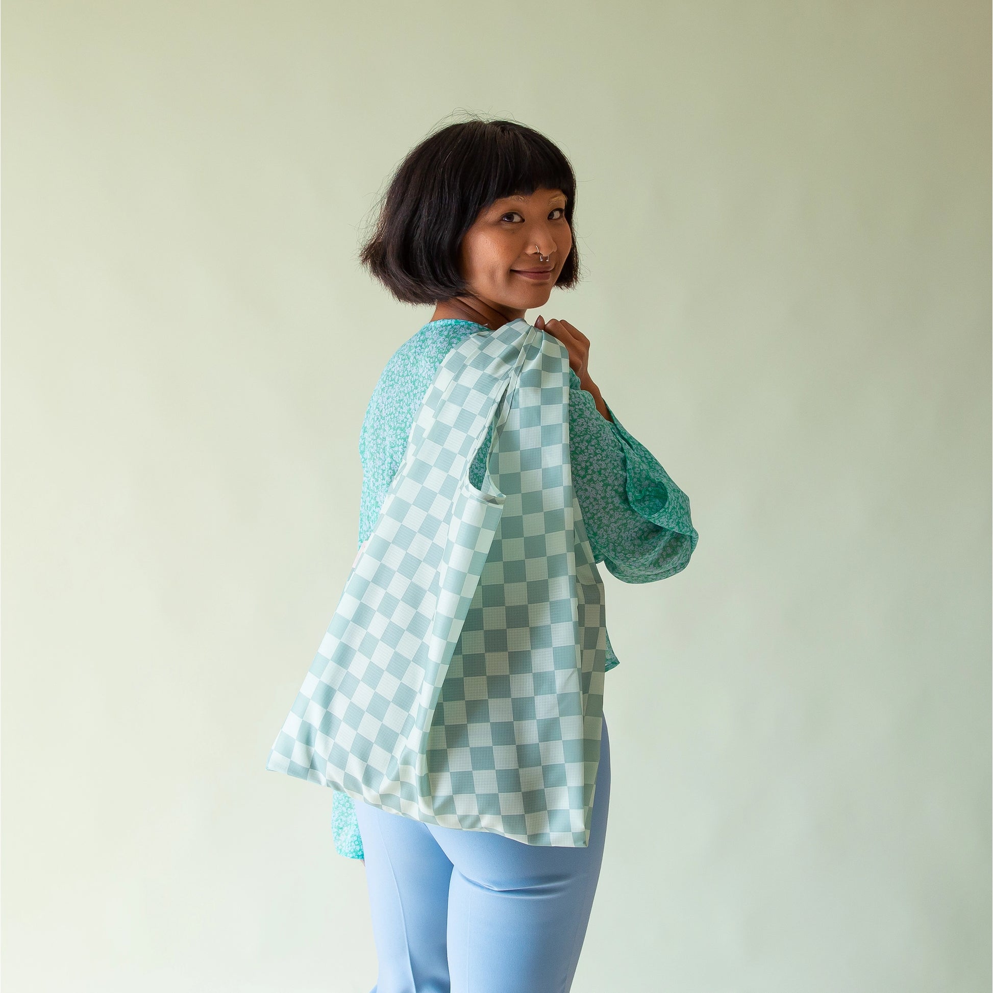 On a light green background is a model holding an aqua colored nylon tote with a checkered print all over.