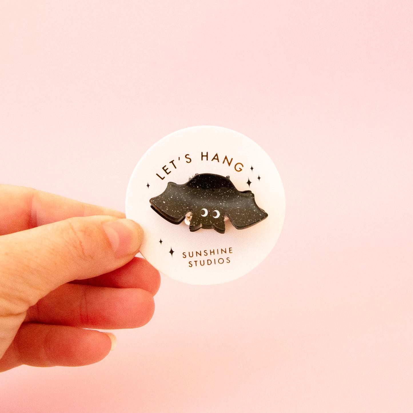 A black speckled bat shaped mini hair clip on a round piece of packaging that reads, 'Let's Hang Sunshine Studios'. 