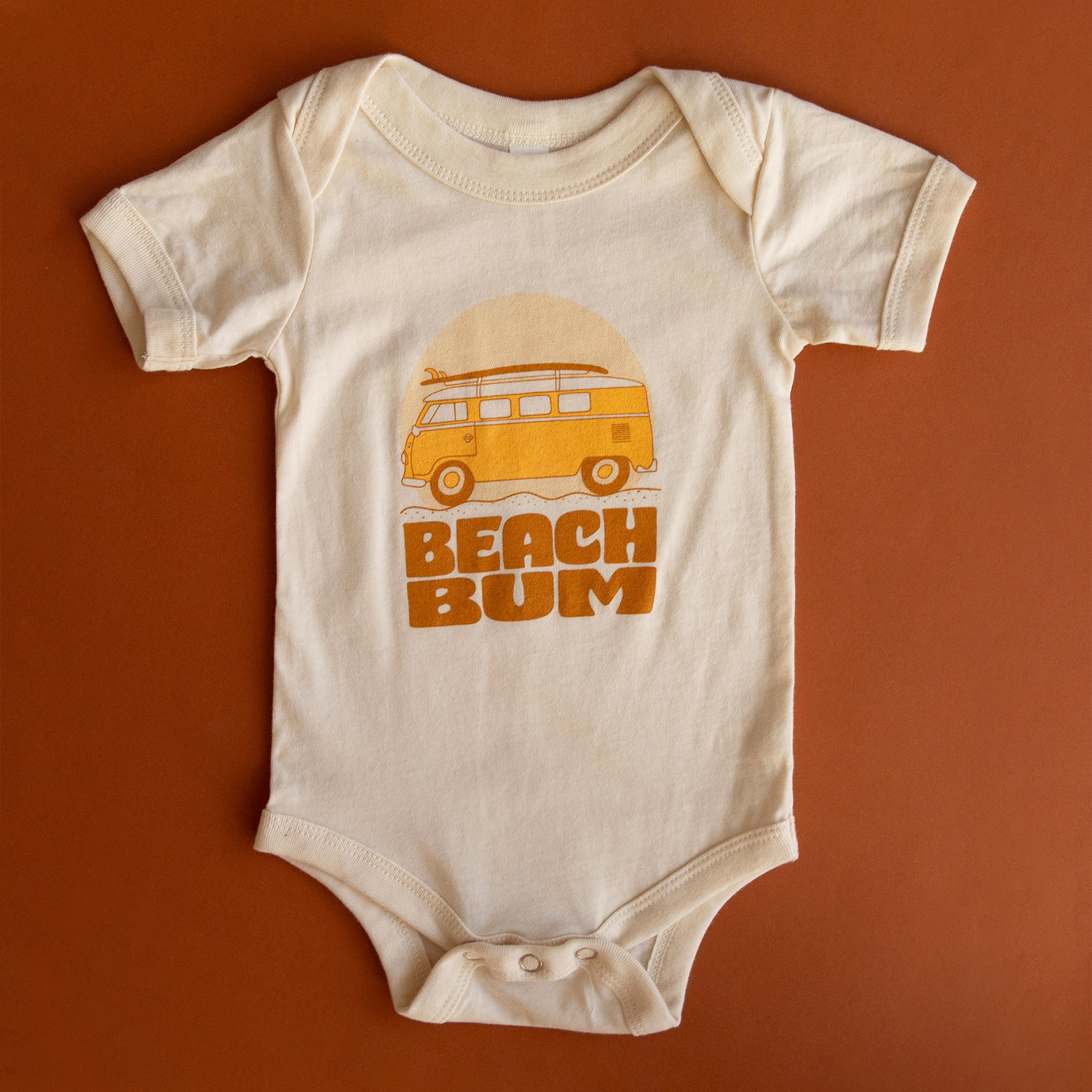 A white cotton onesie with an orange VW bus lined with surfboards and the writing "Beach Bum" in a burnt orange shade.