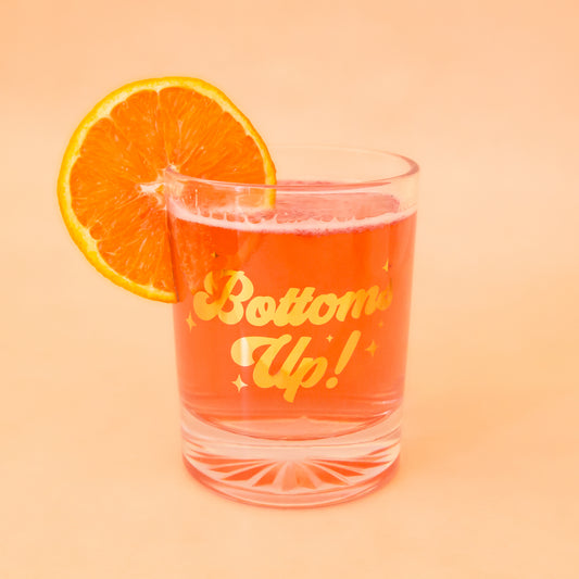Glass tumbler with gold foil writing, spelling"bottoms up!" is shown against a light pink background