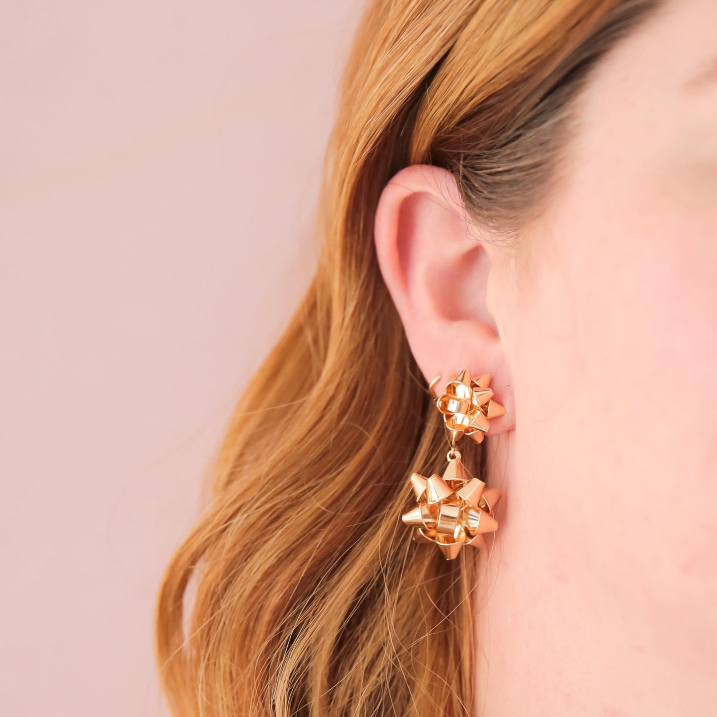 On a peachy background is a model wearing the gold double bow holiday earrings.