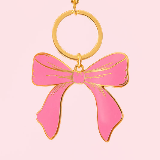 A pink enamel bow keychain with gold hardware hangs on a soft pink background.