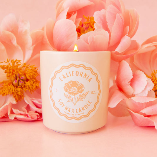 A ceramic candle sits in the middle of image.  The candle has a round white label that reads, "California Soy Wax Candle", in hte background are pink peony flowers.