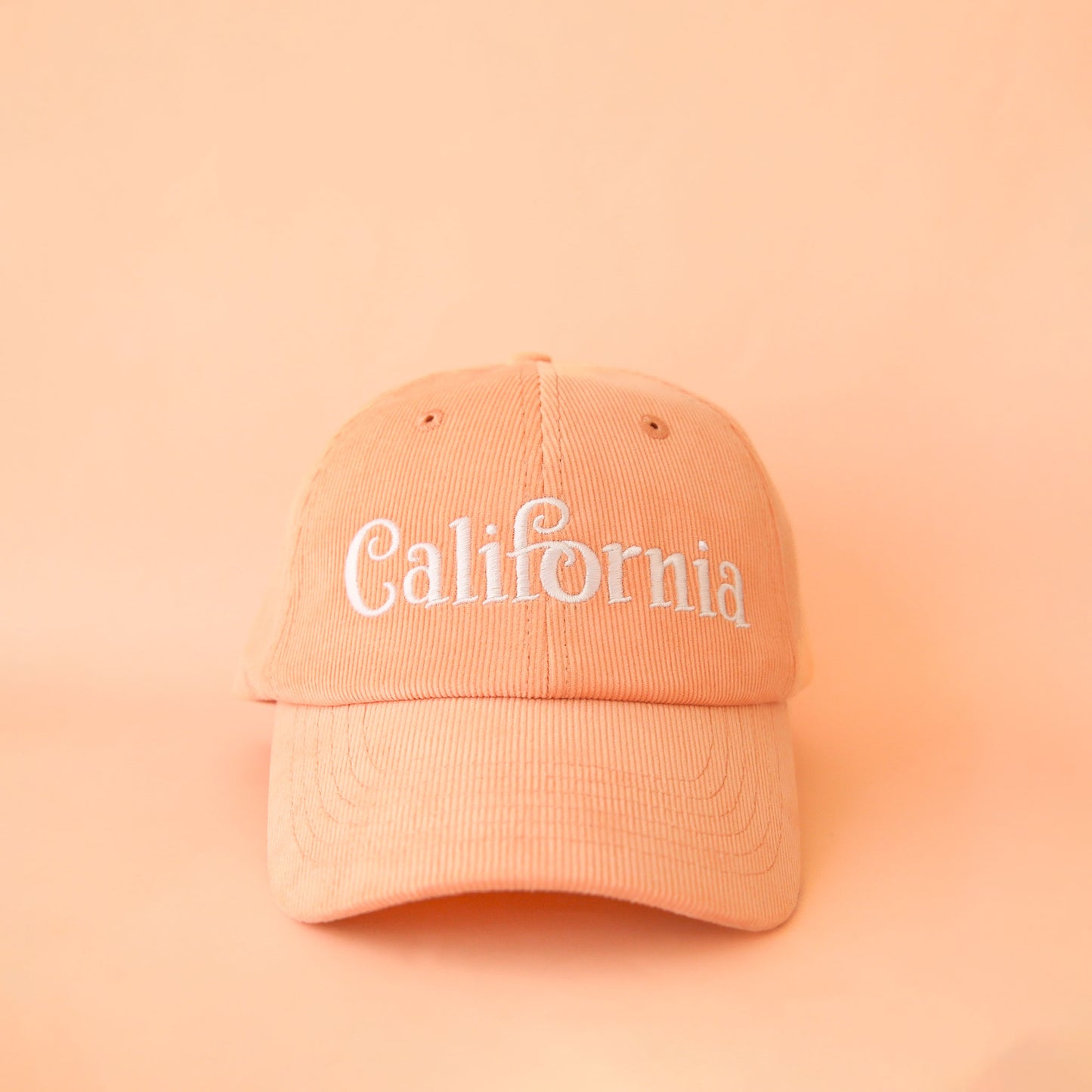 On a peach background is a peach colored baseball hat with a corduroy material and white embroidered text across the front that reads, "California". 