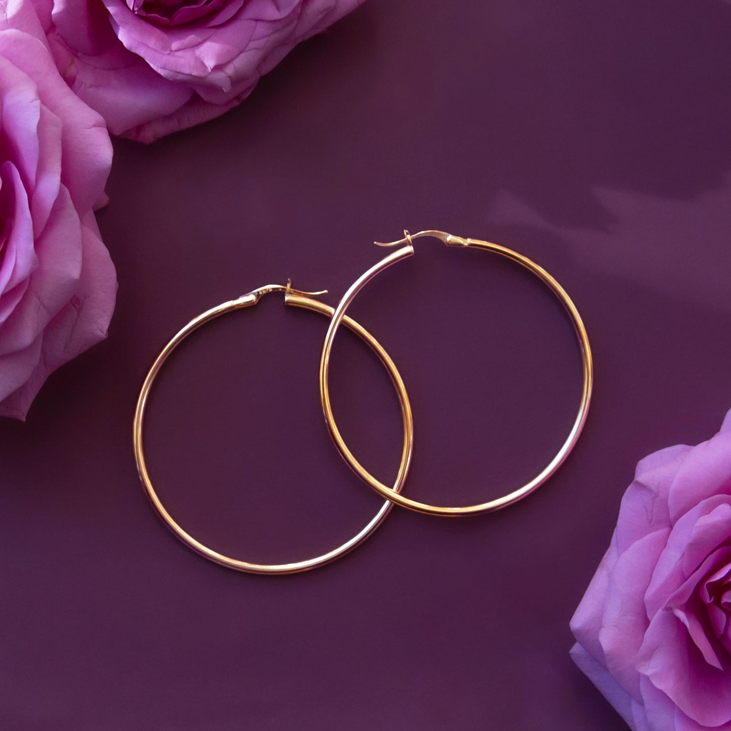 On a pink background is a pair of thin gold hoop earrings next to a stem of pink flowers.